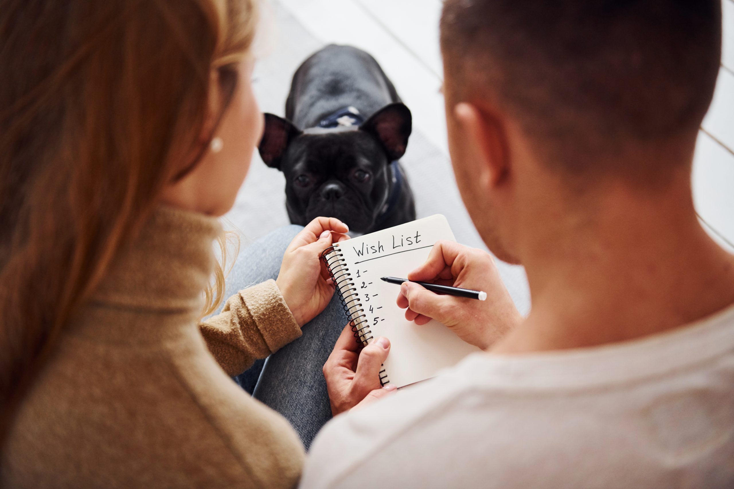 Szczęśliwa młoda para nowoczesny siedzi na kanapie i pisze życzenia w notatniku w domu ze swoim uroczym psem.