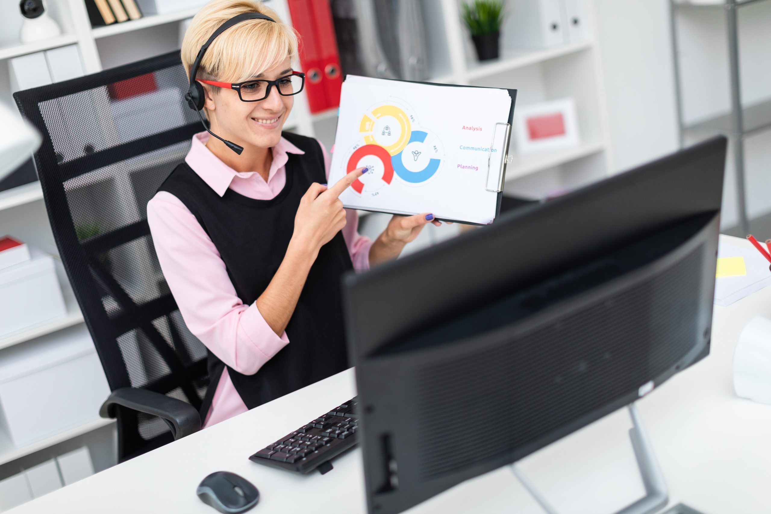 Młoda dziewczyna ze słuchawkami siedzi przy stole komputera i trzyma arkusz na schemacie.