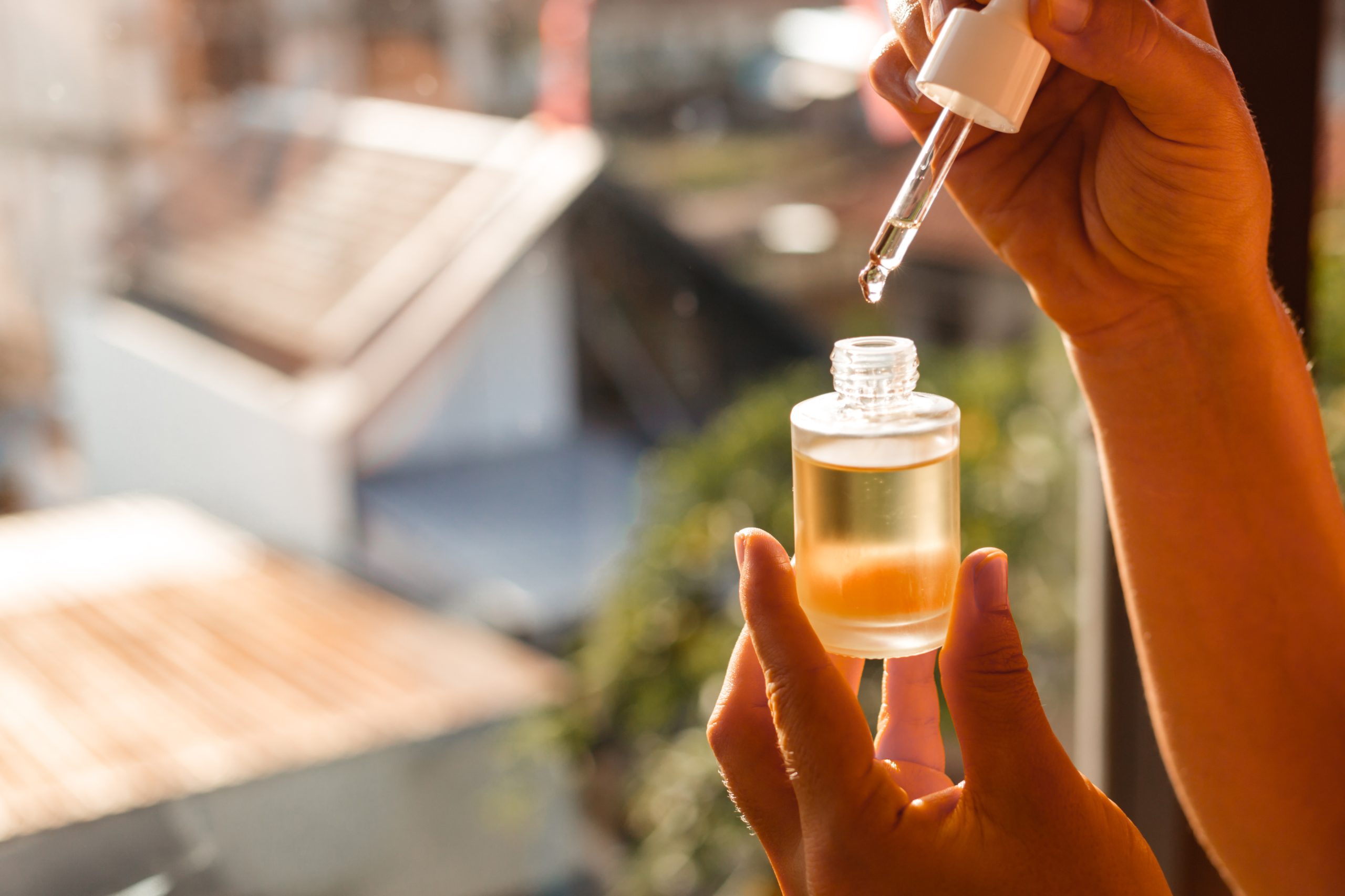 Butelka naturalnego oleju do pielęgnacji twarzy i włosów na tle porannego słońca i promieni. Damskie dłonie trzymają pipetę z naturalnym serum oczyszczającym do twarzy.